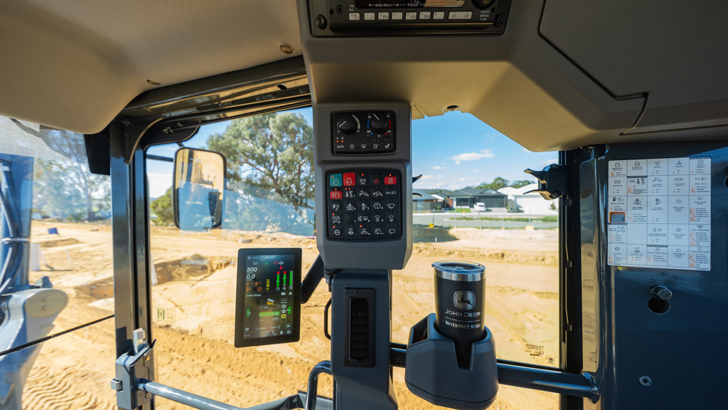 Interior view of a John Deere motor grader cab showing controls, display screen, and work area outside.