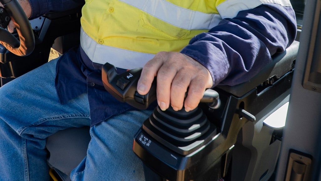 Operator uses a joystick control while seated inside a John Deere motor grader cab