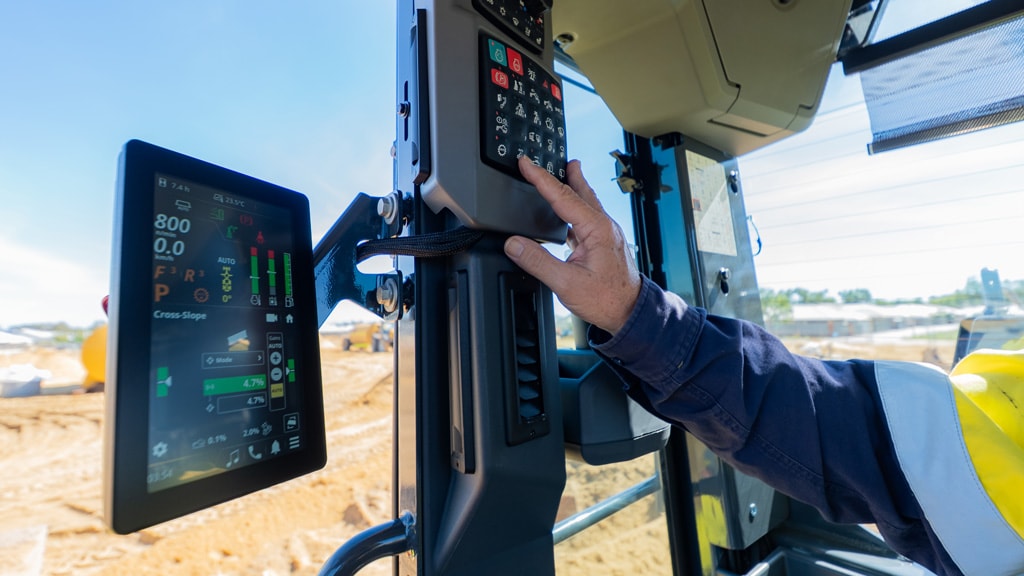 Operator adjusts cab controls beside a display screen in a John Deere motor grader during grading.