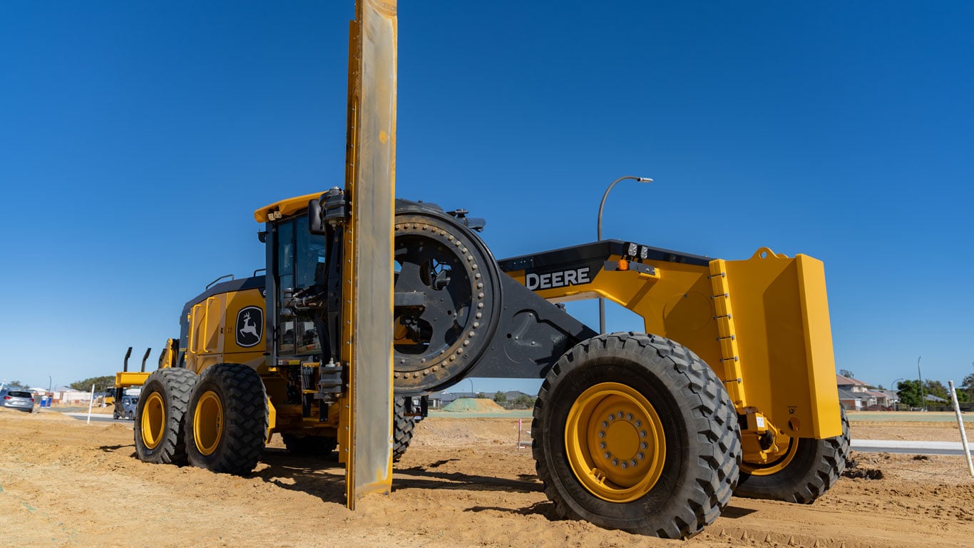 John Deere motor grader works beside a steel beam at a road construction site.
