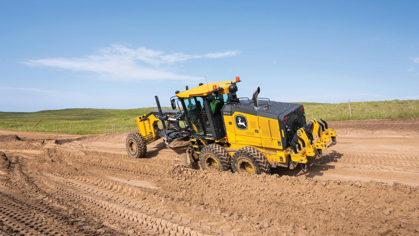 John Deere motor grader levels a dirt road on a construction site under a clear blue sky