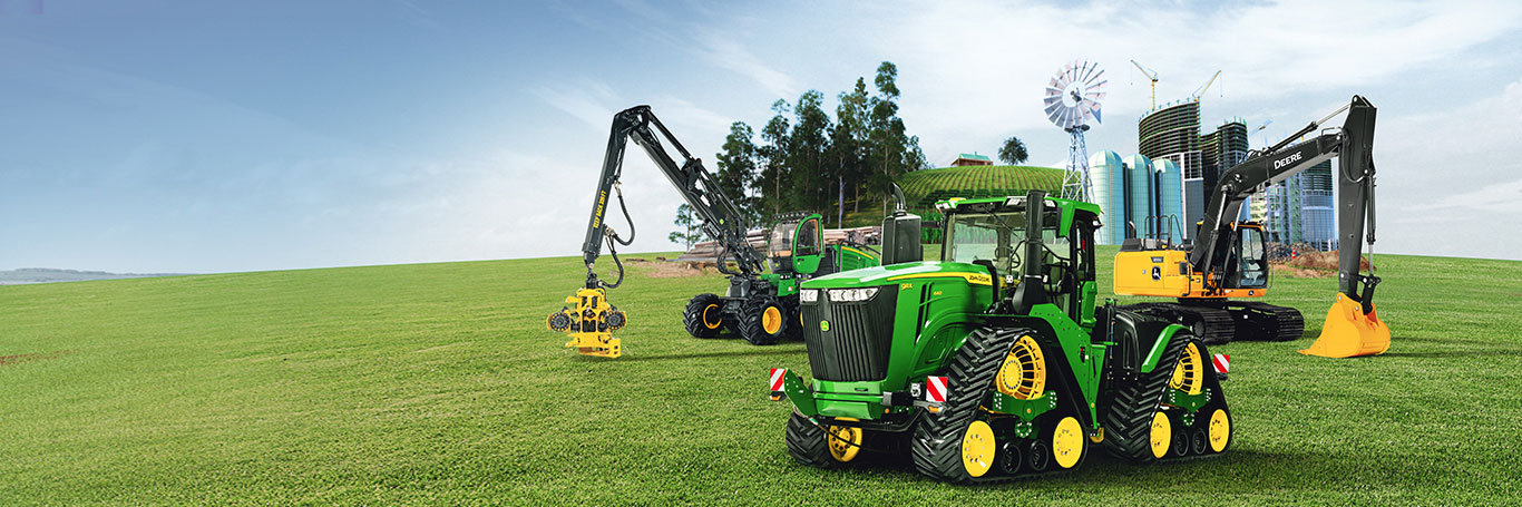 Three pieces of John Deere agriculture, construction and forestry equipment in a field foreground with a farm, forest and large building construction site in the background.