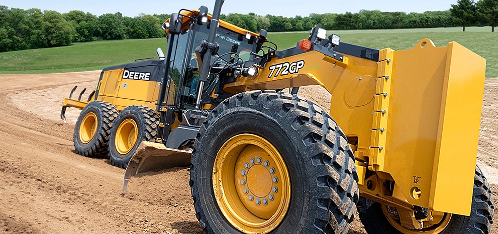 A side shot of a 772gp Motor Grader on a dirt track by the forest on a sunny day