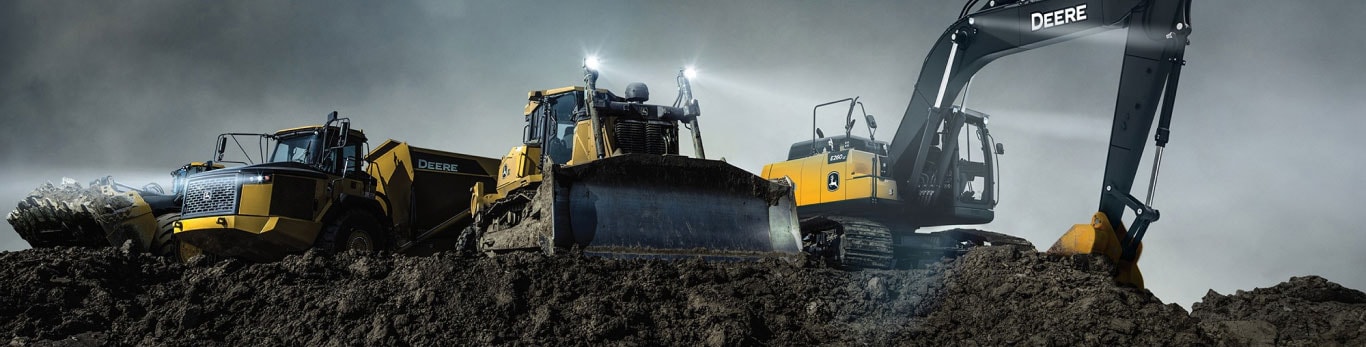A line up of John Deere construction equipment including an Articulated Dump Truck, Dozer and Excavator, pictured against a dark and stomy sky on a mound of earth