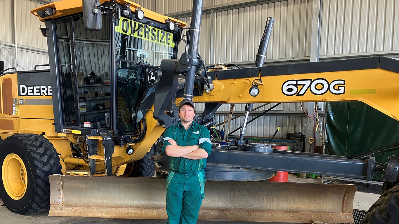 Ben Mummery with arms crossed leaning on a 670G motor grader