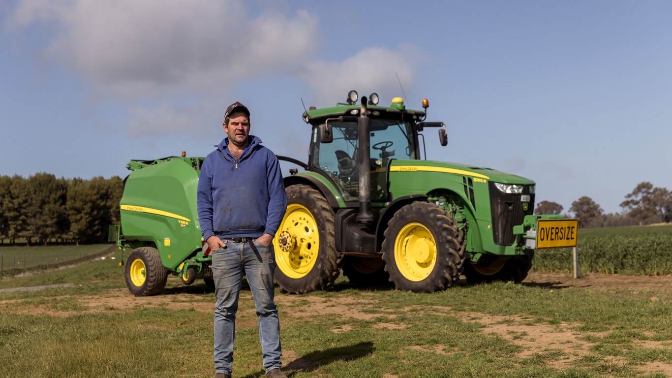 Farmer standing in front of tractor with hands in its pockets on an empty grass field.