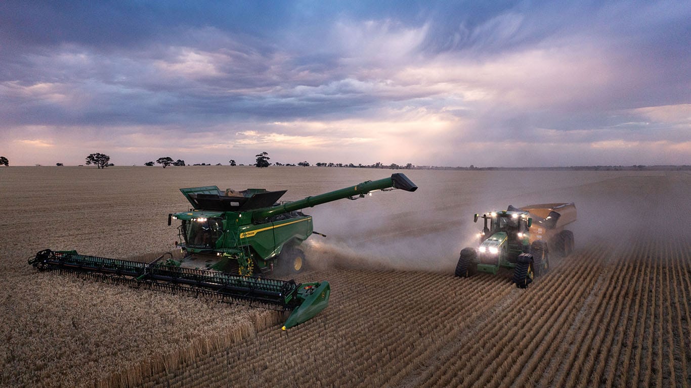 A John Deere Combine Harvester and tractor with chaser bin in a wheat paddock at dusk