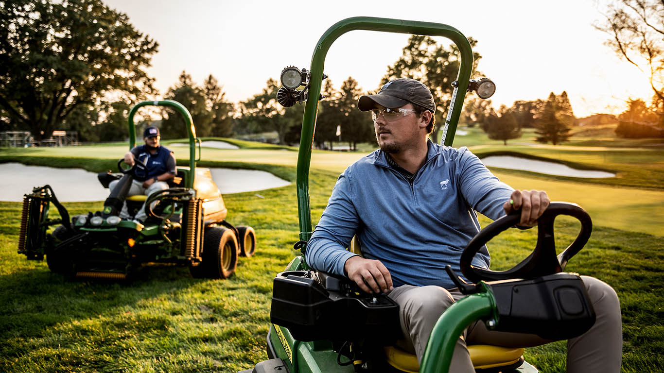 Two Men each sitting on a golf mower, looking out onto the fairway