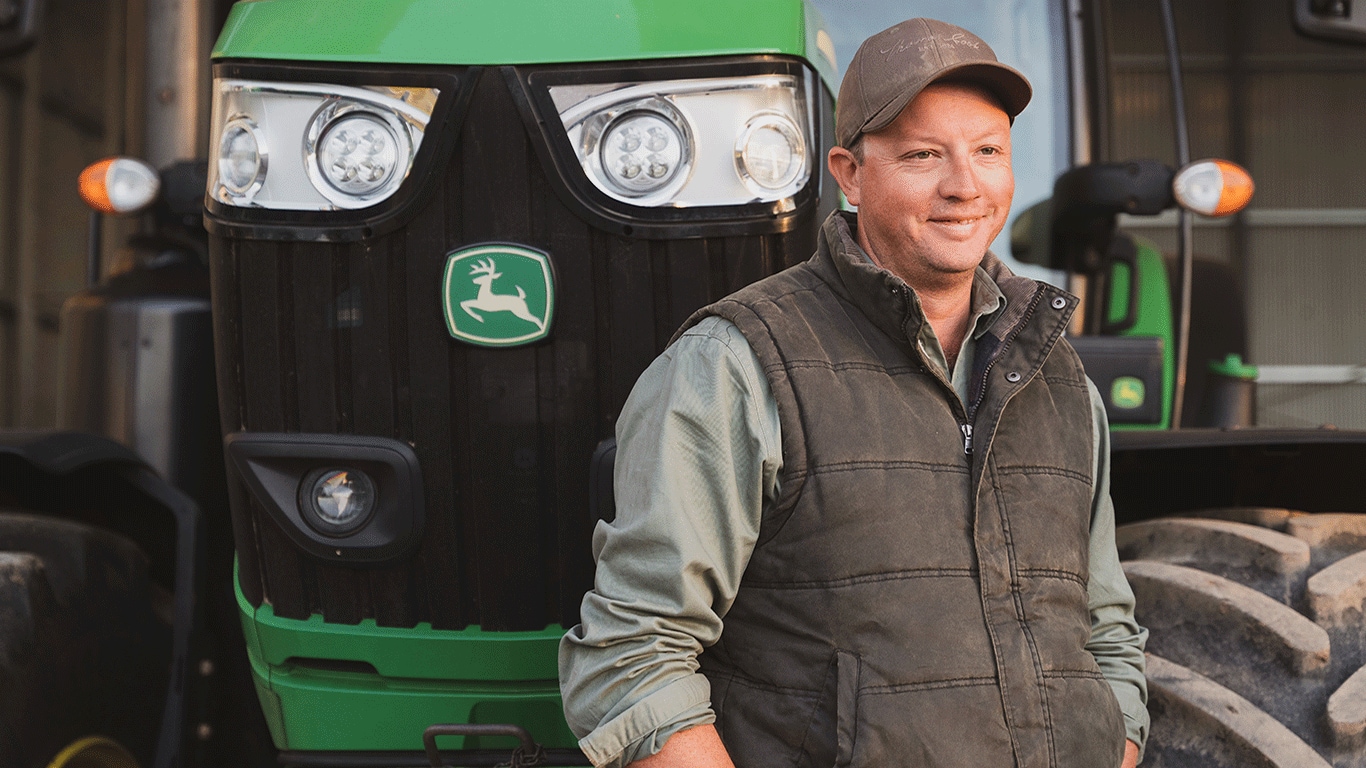 Farmer Richard Stecher stands in front of one of his John Deere tractors. He's smiling and looking off in the distance.