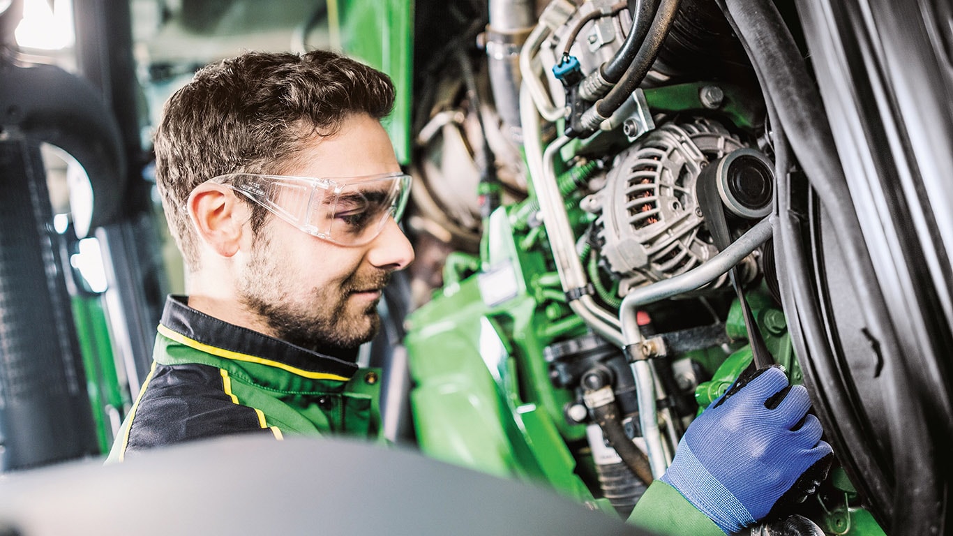 A John Deere technician is working on a pristine looking engine.  He's got his PPE goggles and gloves on and he's making adjustments to a belt.