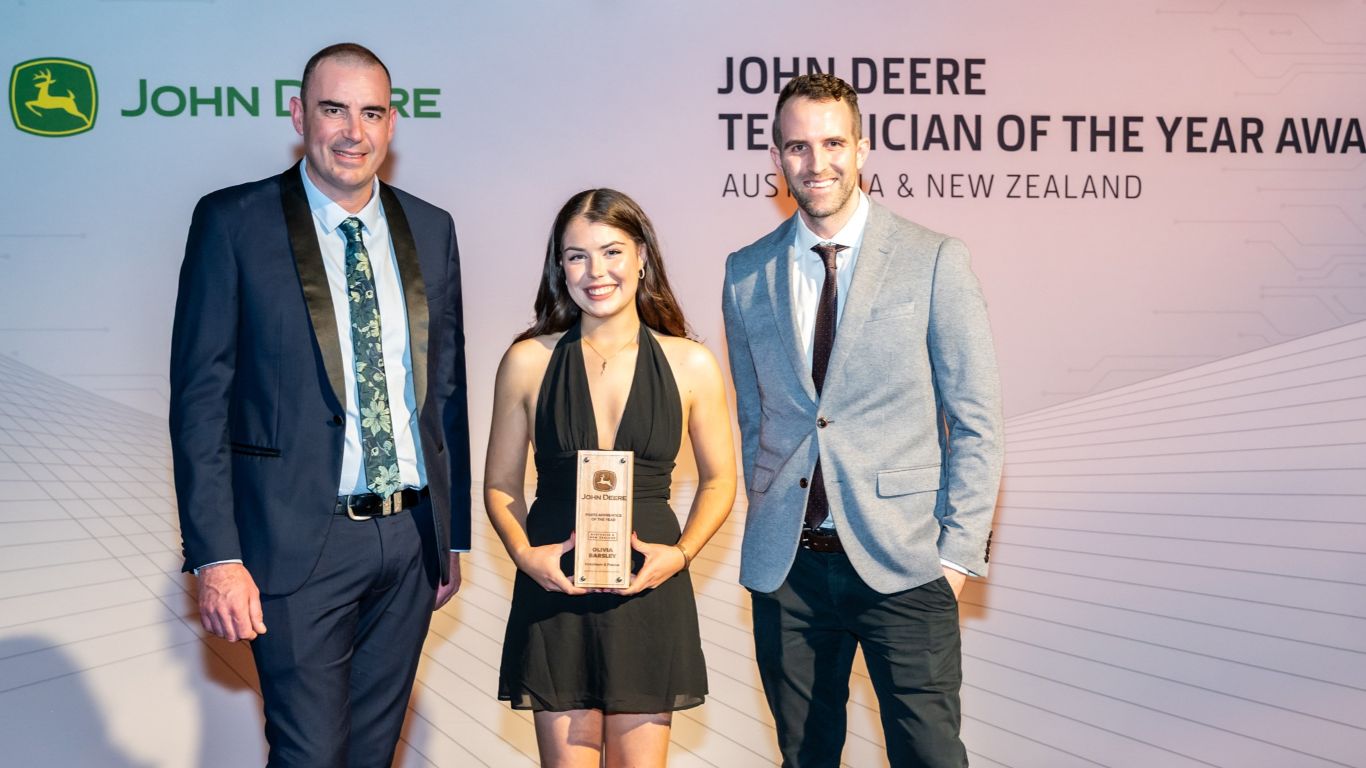 A lady holding a wooden trophy stands in front of a media wall with a man on either side.