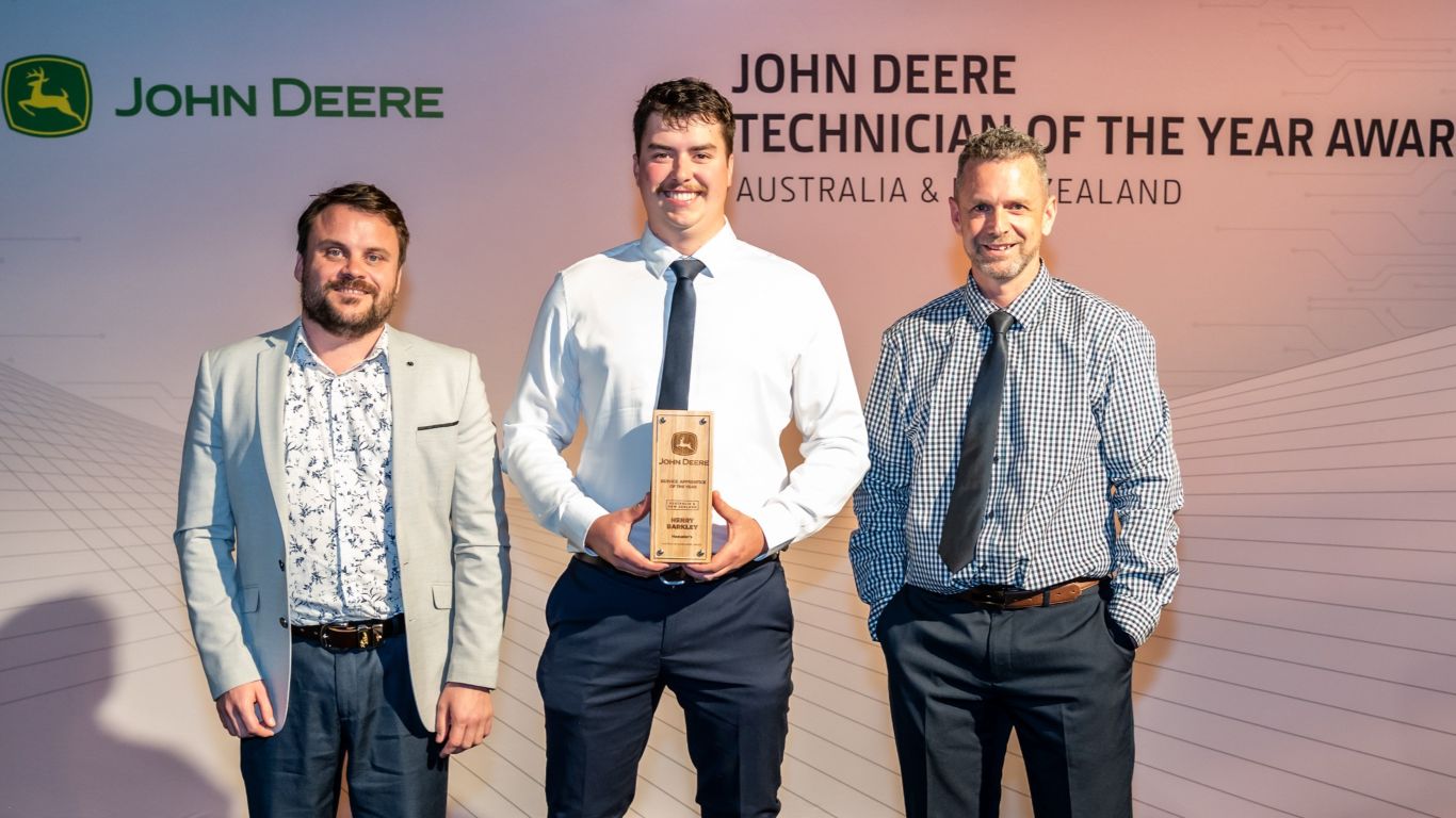 Three men standing in front of a media wall with the man in the centre holding a trophy.
