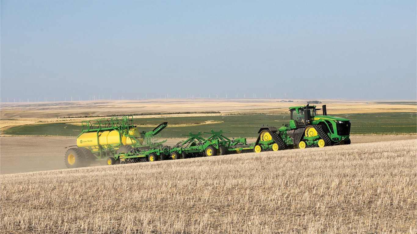 John&nbsp;Deere 9RX Tractor with P600 Precision Air Hoe Drill and C-Series Air Cart in paddock with wind turbines in the background and blue sky.