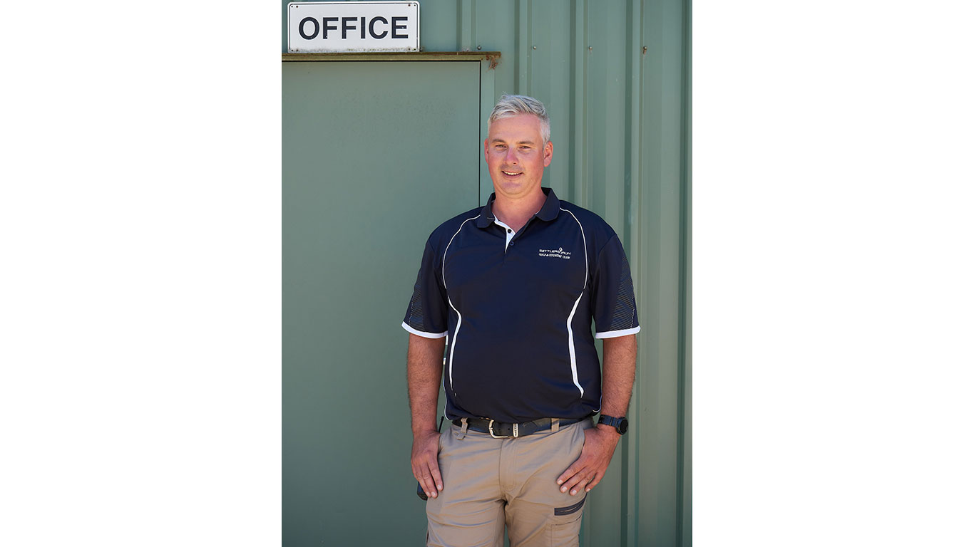 A man with short grey hair stands in front of a green metal building with a sign that reads "OFFICE." He wears a navy blue polo shirt with white piping and beige cargo pants. He has a relaxed posture, with one hand resting on his thigh and the other at his side, smiling at the camera.