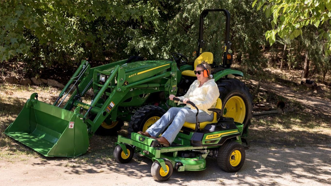 A woman in light yellow blouse is on a John&nbsp;Deere ride-on mower with a tractor and trees behind them.