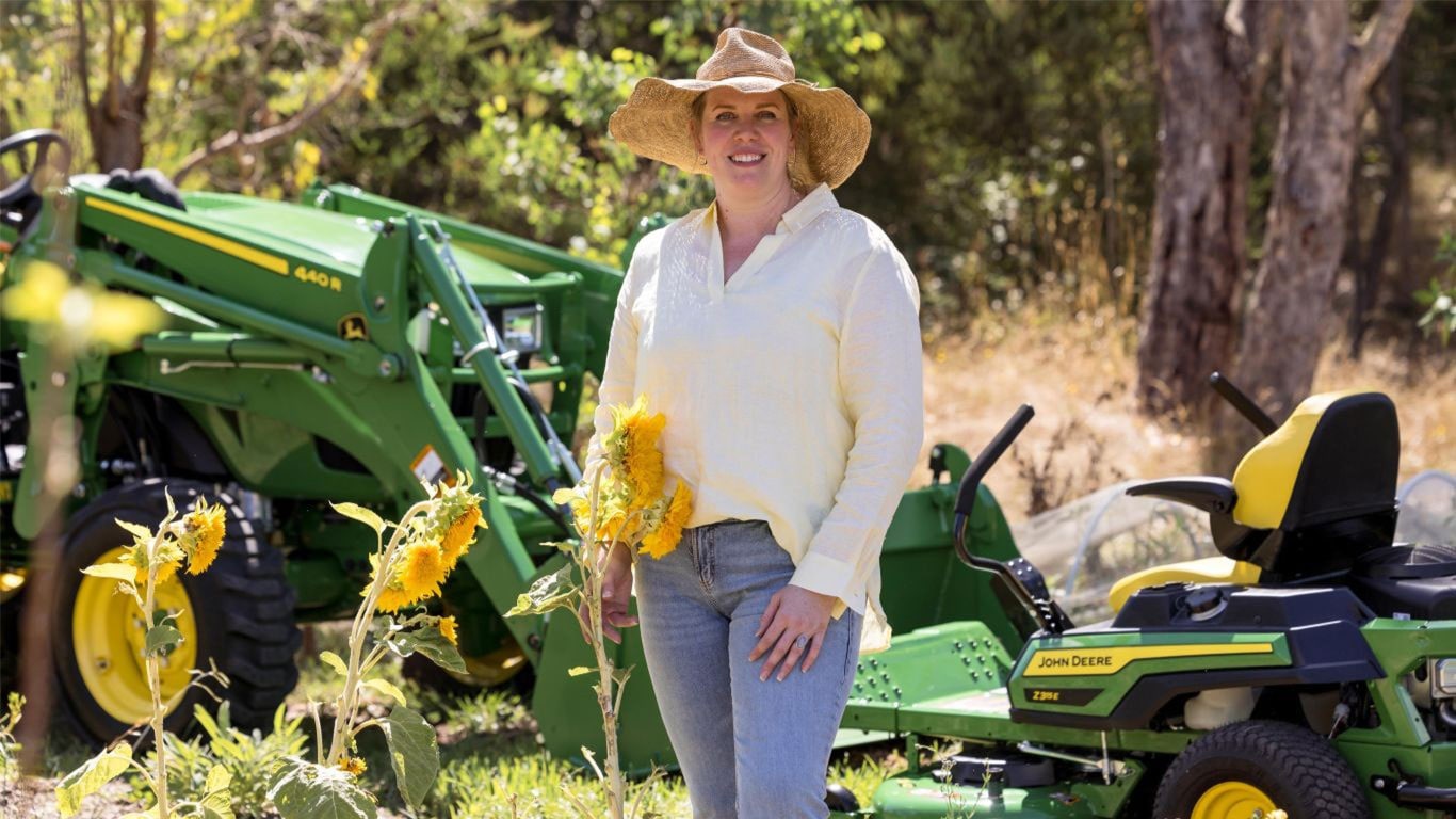 A woman in a straw hat and light yellow blouse stands in a garden holding a sunflower, with John&nbsp;Deere equipment and trees in the background.