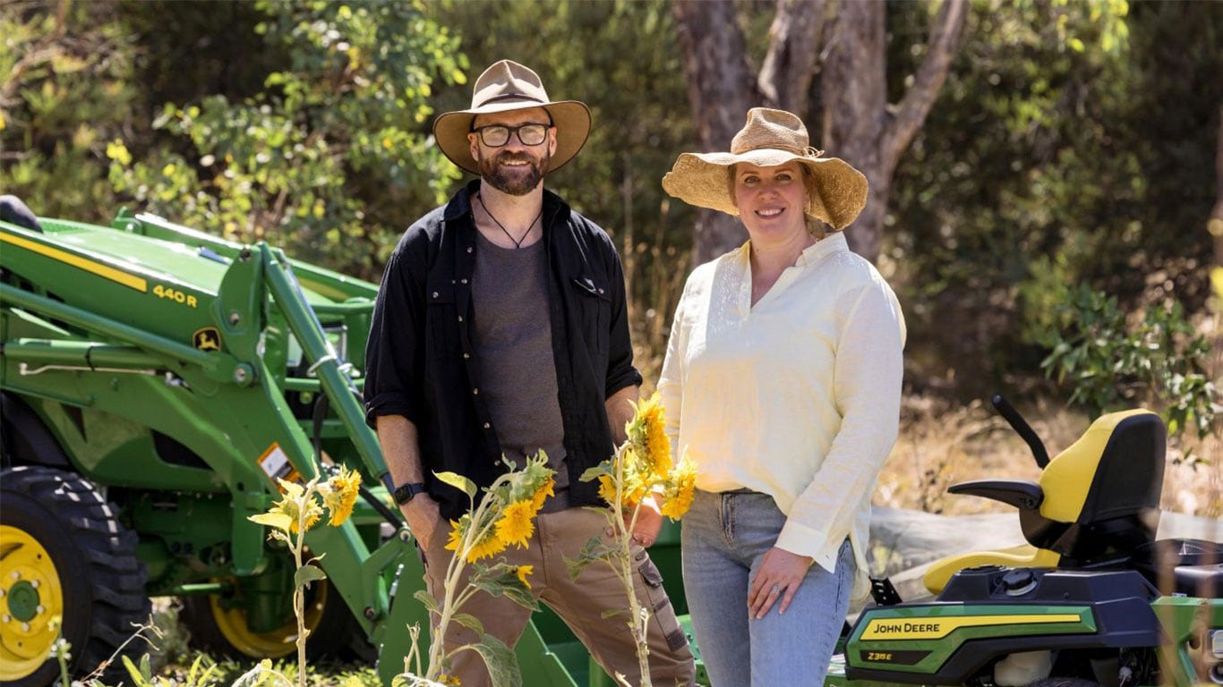 A man and woman in wide-brimmed hats stand among sunflowers, smiling, with John&nbsp;Deere equipment and trees in the background.