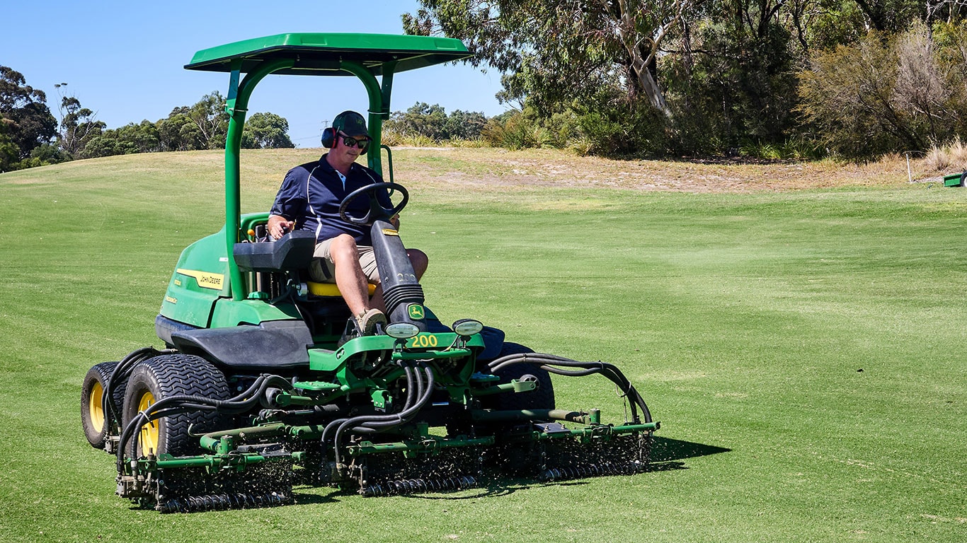 A man wearing a navy blue polo shirt, sunglasses, gap and earmuffs operates a large green mower.