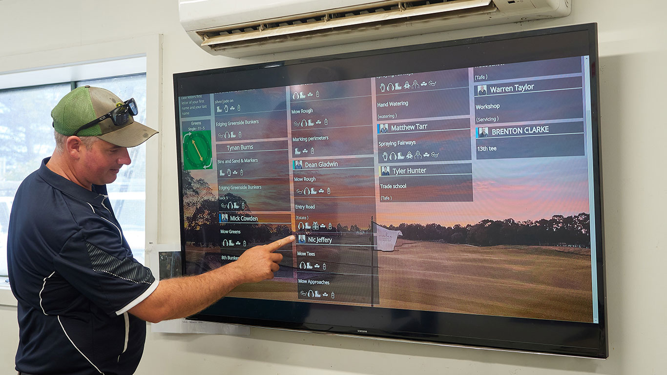 A man in a navy blue polo shirt and a green cap stands inside an office, pointing at a large wall-mounted screen displaying a golf course management system. The screen shows various tasks, staff names, and a scenic golf course image as the background.