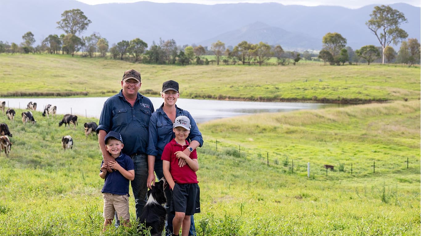 A family of four with their dog are standing in a green pasture with cows and hills in the background.