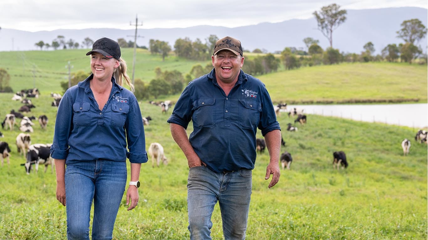 A male and a female are smiling while walking through a green pasture with grazing cows and hills in the background.