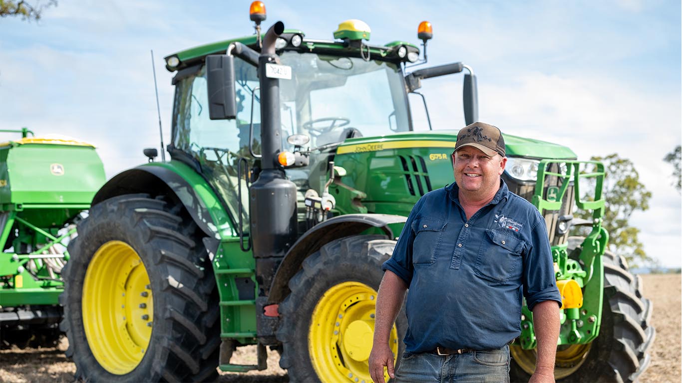 A man in a blue shirt is standing in front of a John&nbsp;Deere tractor.