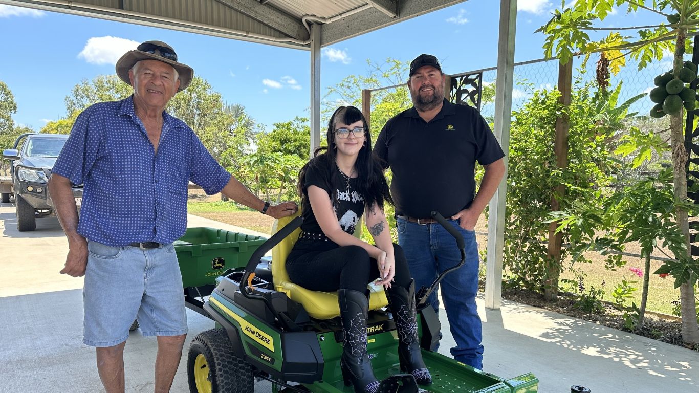 An elderly man, a young woman, and another man stand together under a carport with a John Deere zero-turn mower. The young woman is seated on the mower, while the older man rests his hand on the seat and the third man stands beside them.