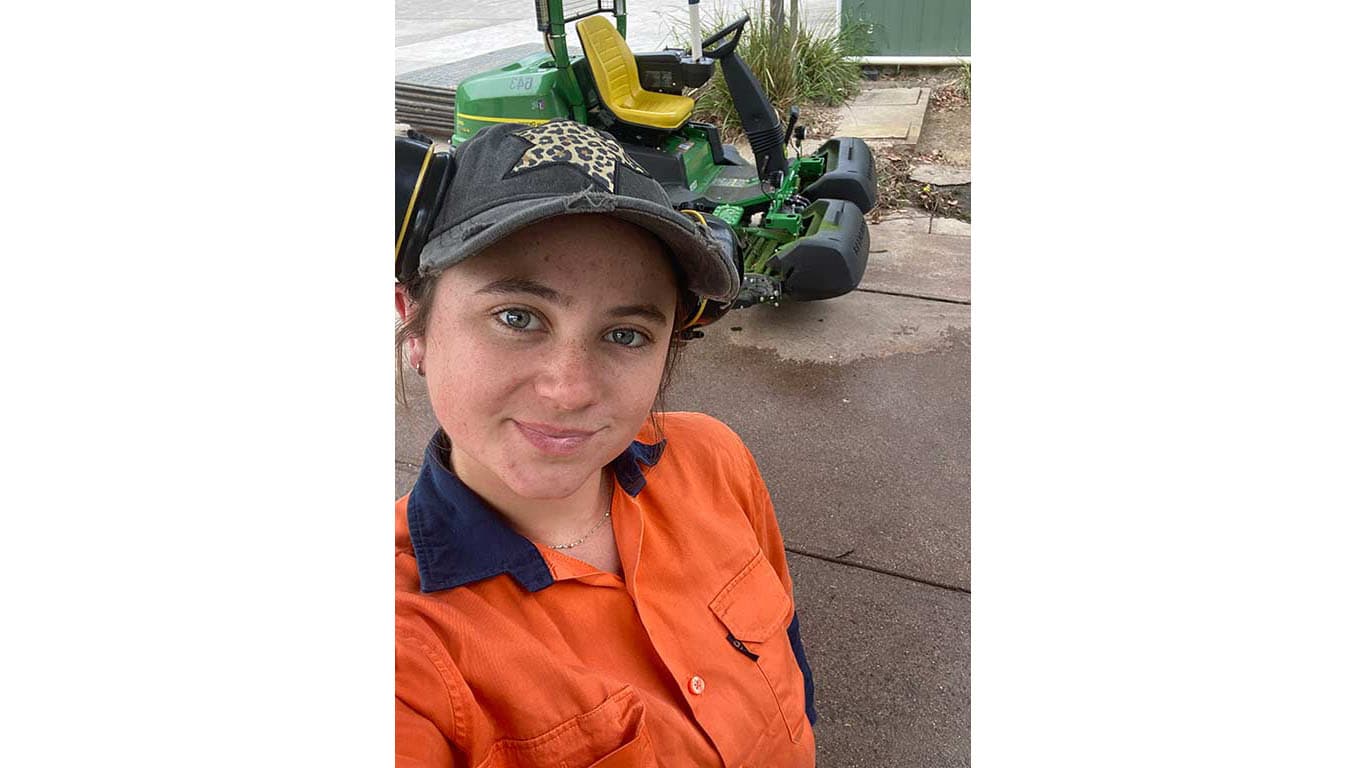 Ella Arter smiling at camera with John Deere fairway mower in background