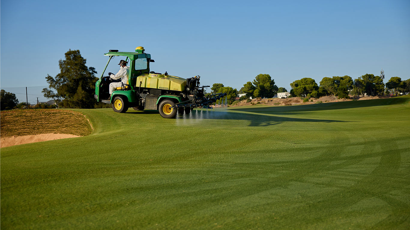 A worker in a white protective suit and mask operating a John Deere 2030A GPS PrecisionSprayer on a golf course fairway.