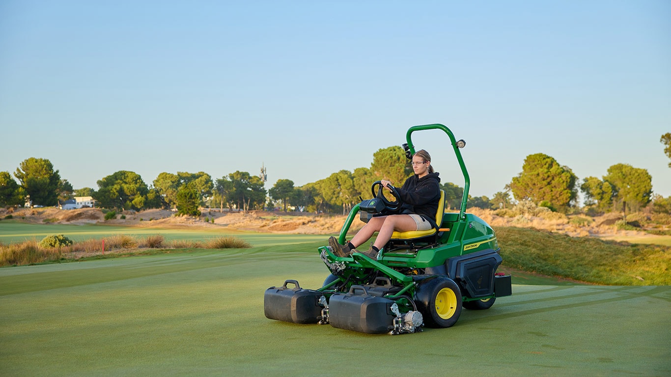 A young woman operating a John Deere 2750 E-Cut Hybrid Triplex mower on a golf course green.