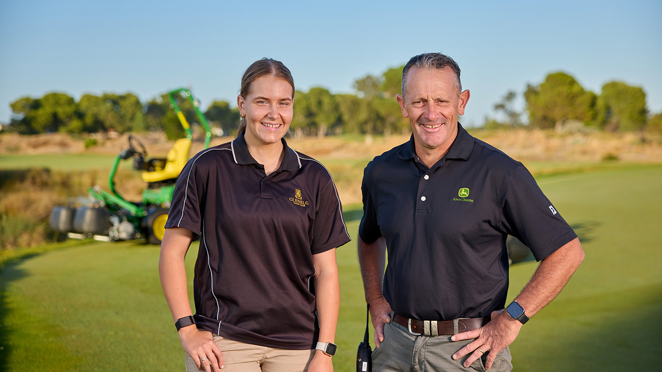 A smiling man and young woman standing on a golf course with a John Deere 2750 E-Cut Hybrid Triplex mower blurred in the background.