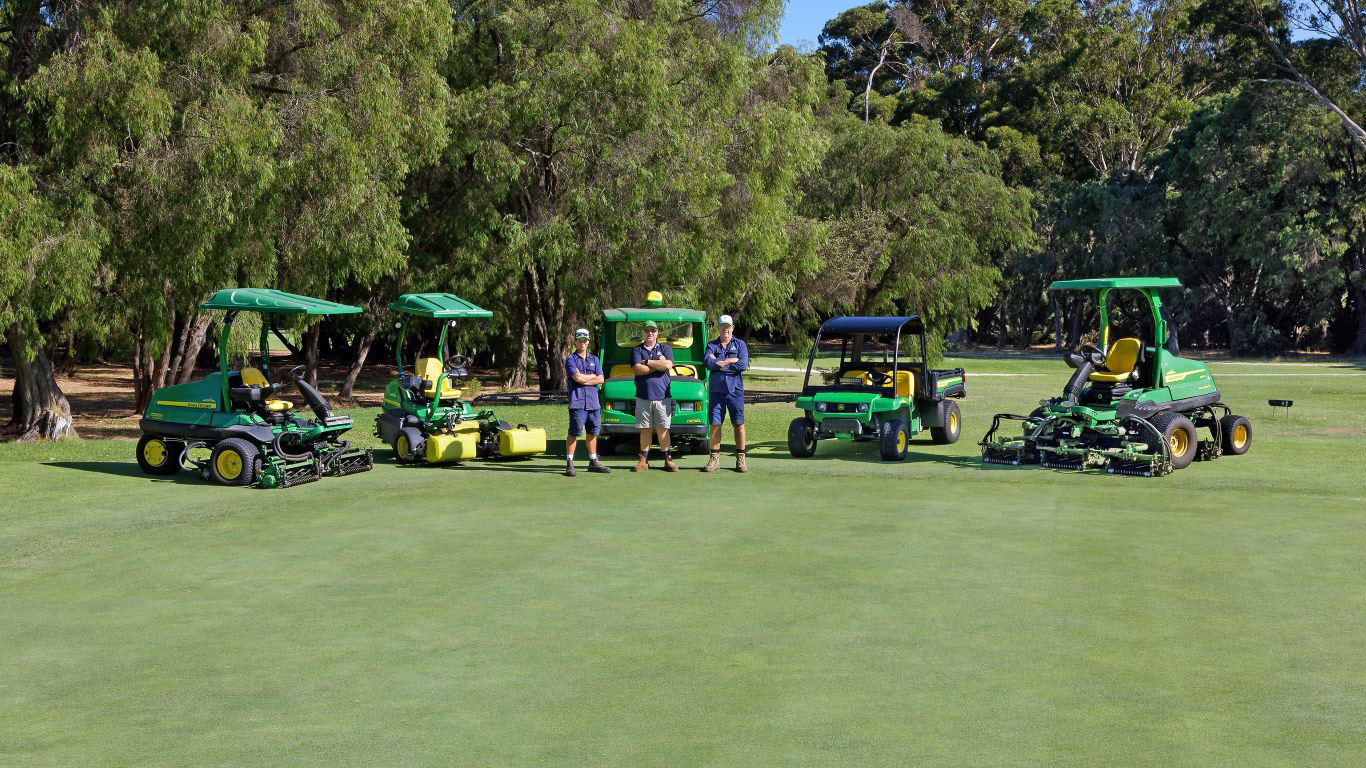 Three people wearing blue shirts, standing on a golf course with five different types of green mowing equipment behind them.