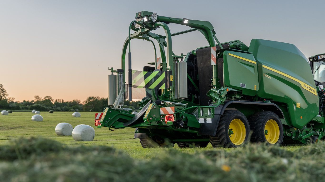 A John Deere baler in operation in a field with wrapped bales.