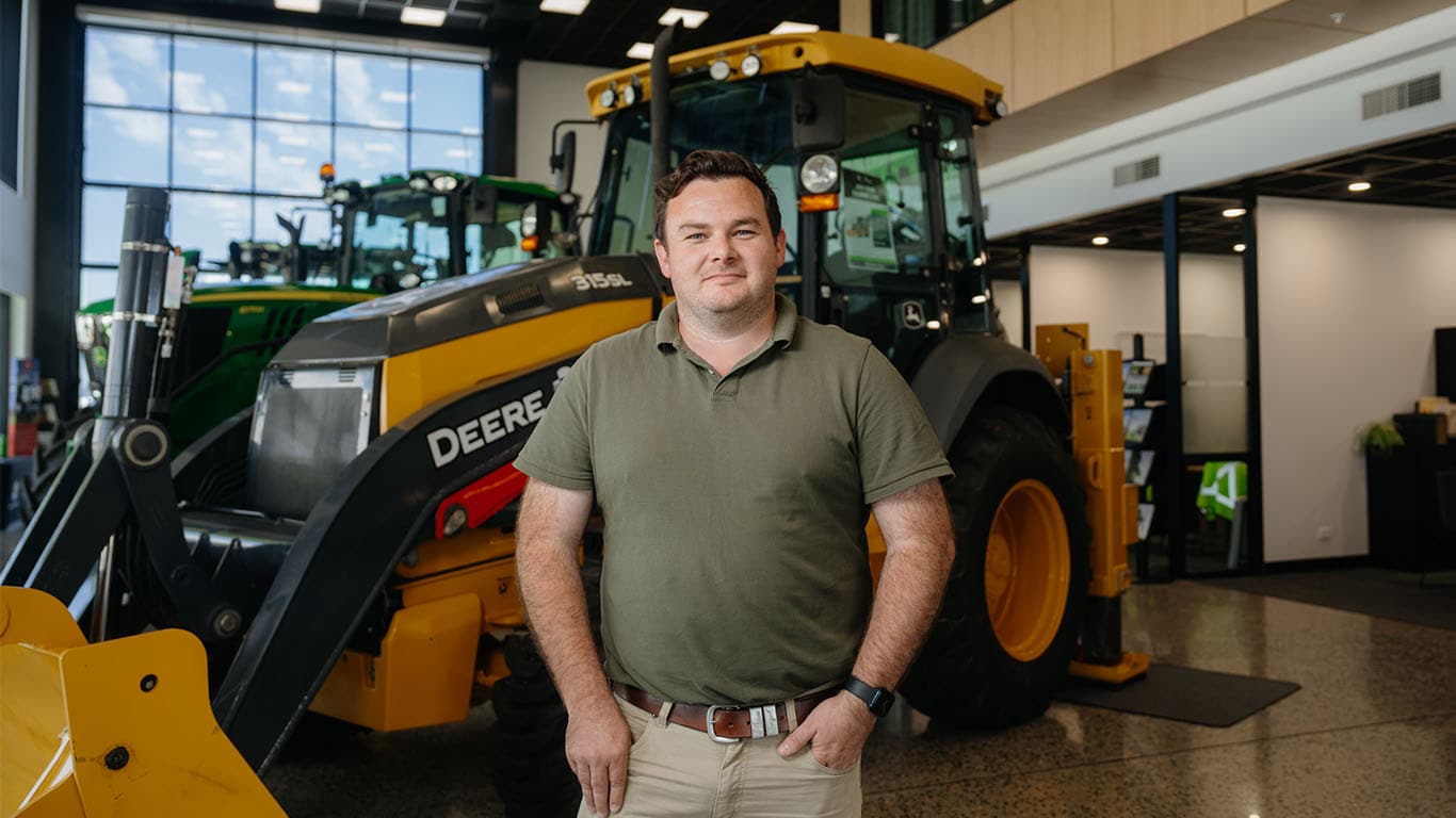 A man stands in front of a yellow John Deere tractor inside a showroom, wearing a green polo shirt and beige pants, with tractors visible in the background under bright indoor lighting.