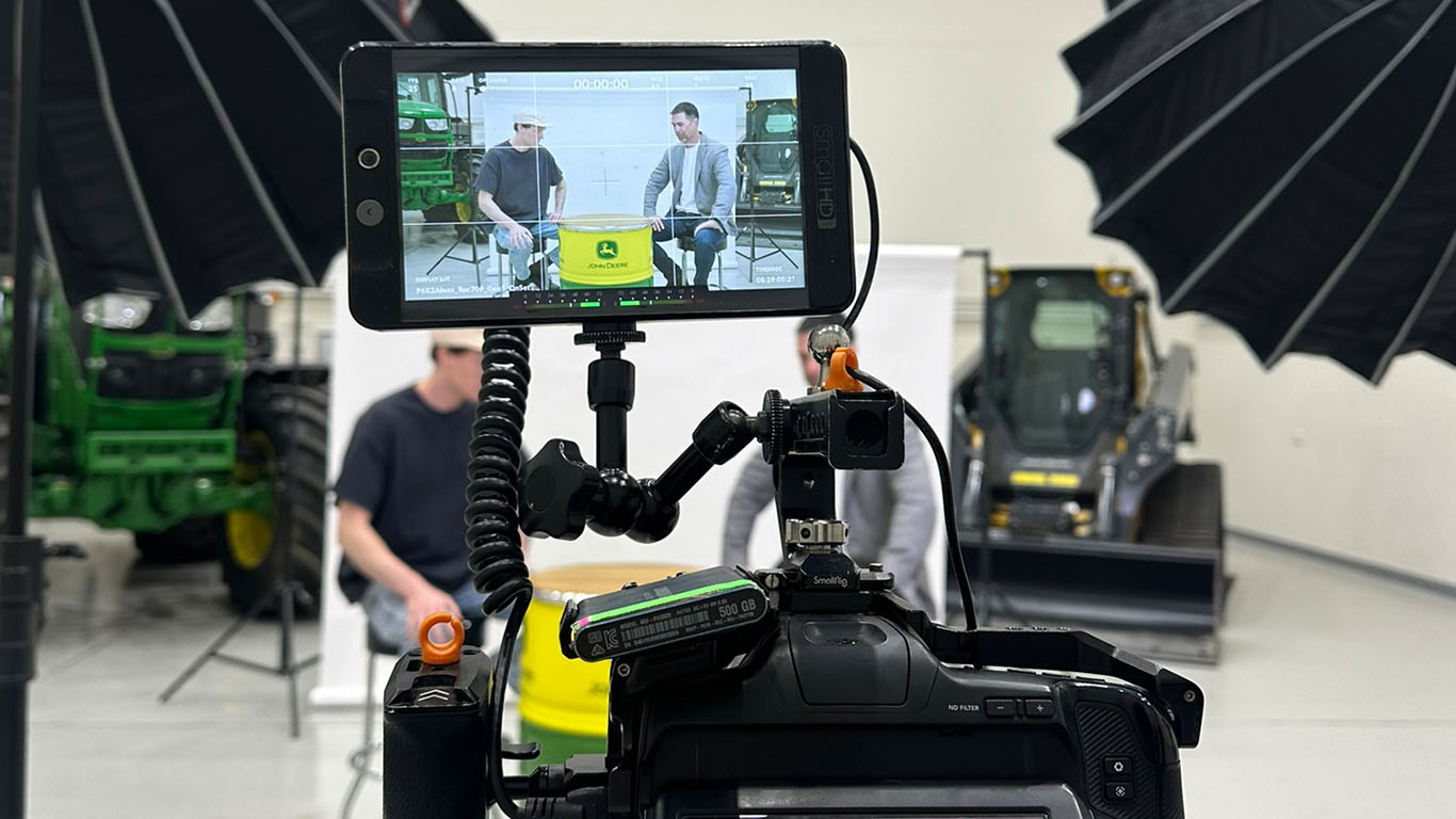 A behind-the-scenes look at a video production setup featuring a camera screen displaying two seated individuals during an interview, surrounded by professional lighting equipment in a warehouse setting with John Deere machinery in the background.