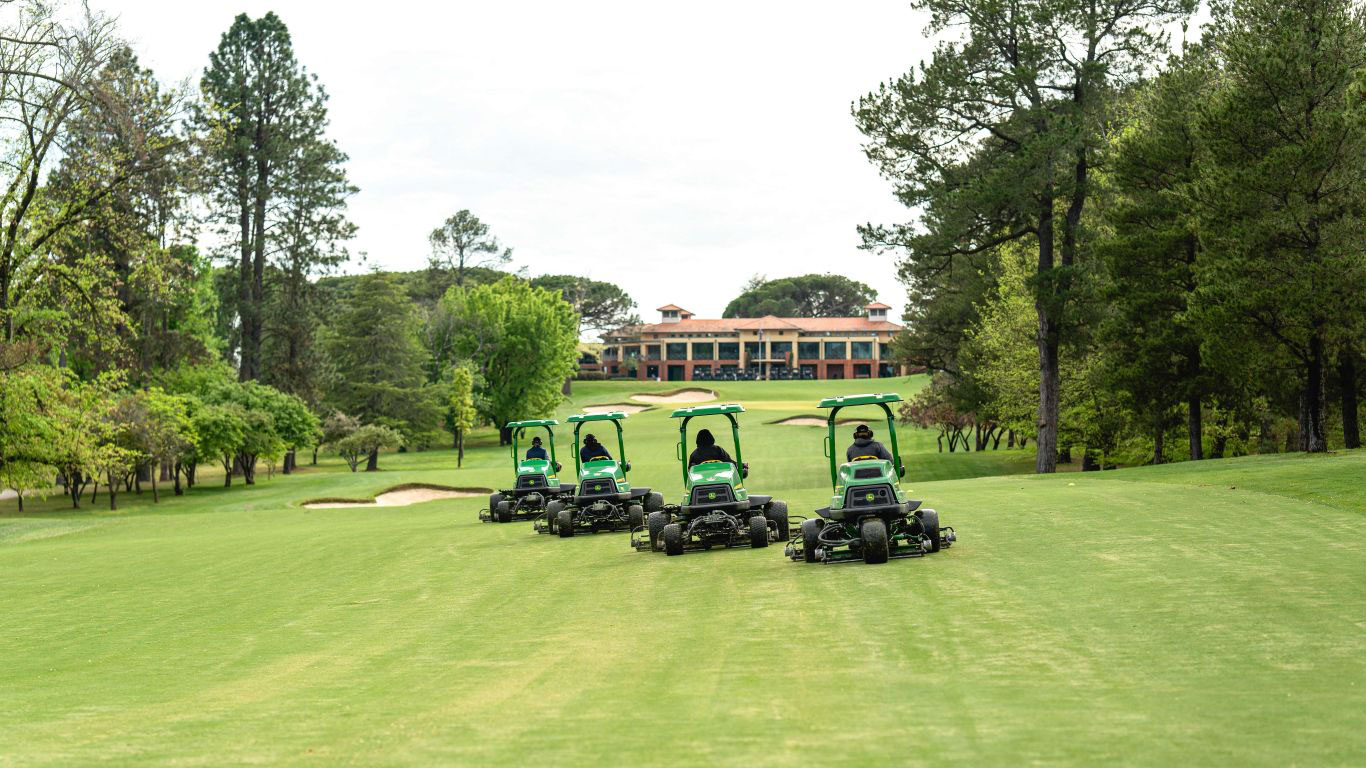 Four green fairway mowers on a golf course fairway flanked by trees and a building is in the background.