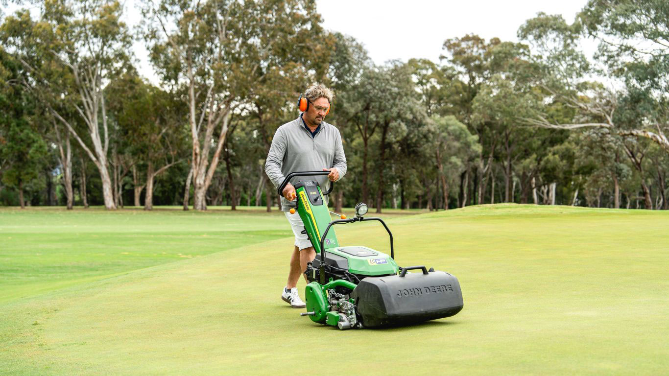 A man with orange earmuffs is pushing a greens mower on a golf course with trees in the background.