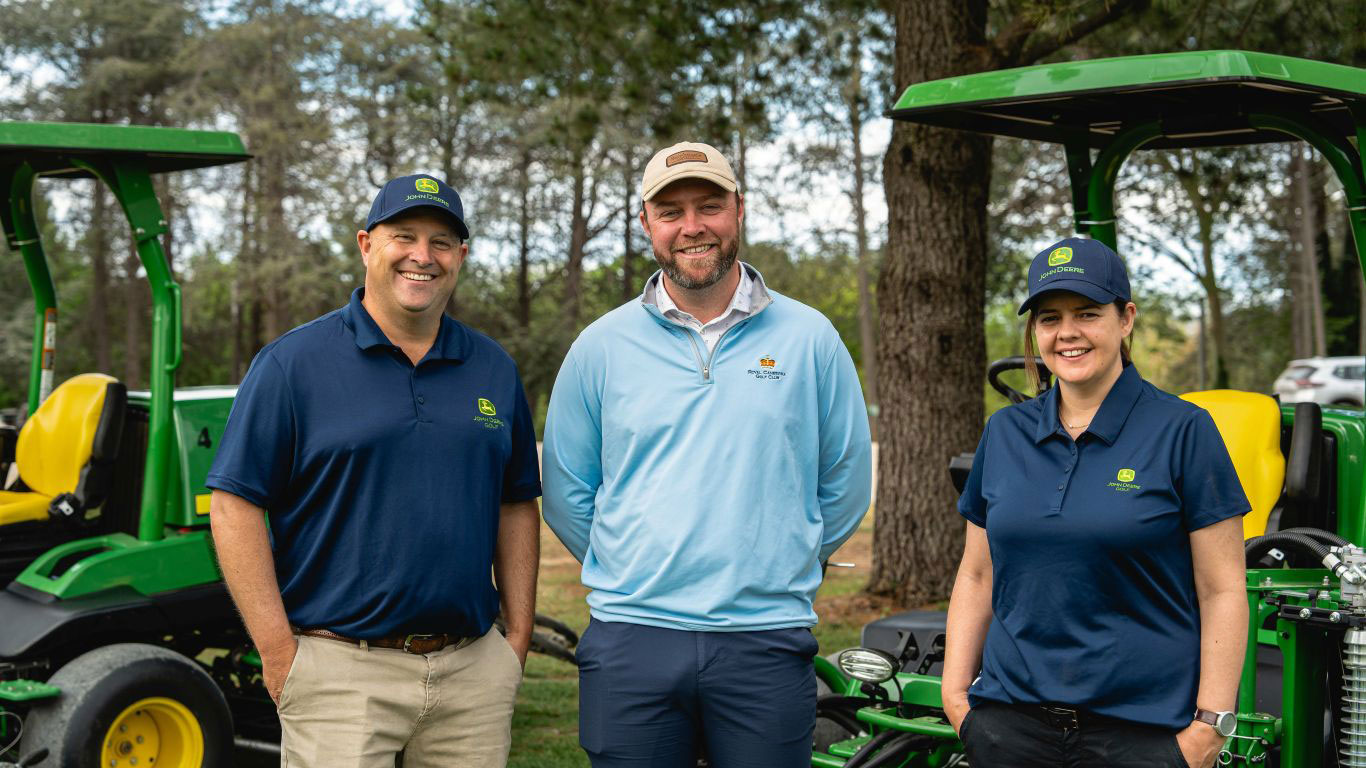 A man in light blue jumper man is standing next to a man wearing a navy blue polo shirt and a lady wearing a navy-blue polo shirt. There are trees behind them and John Deere turf equipment.