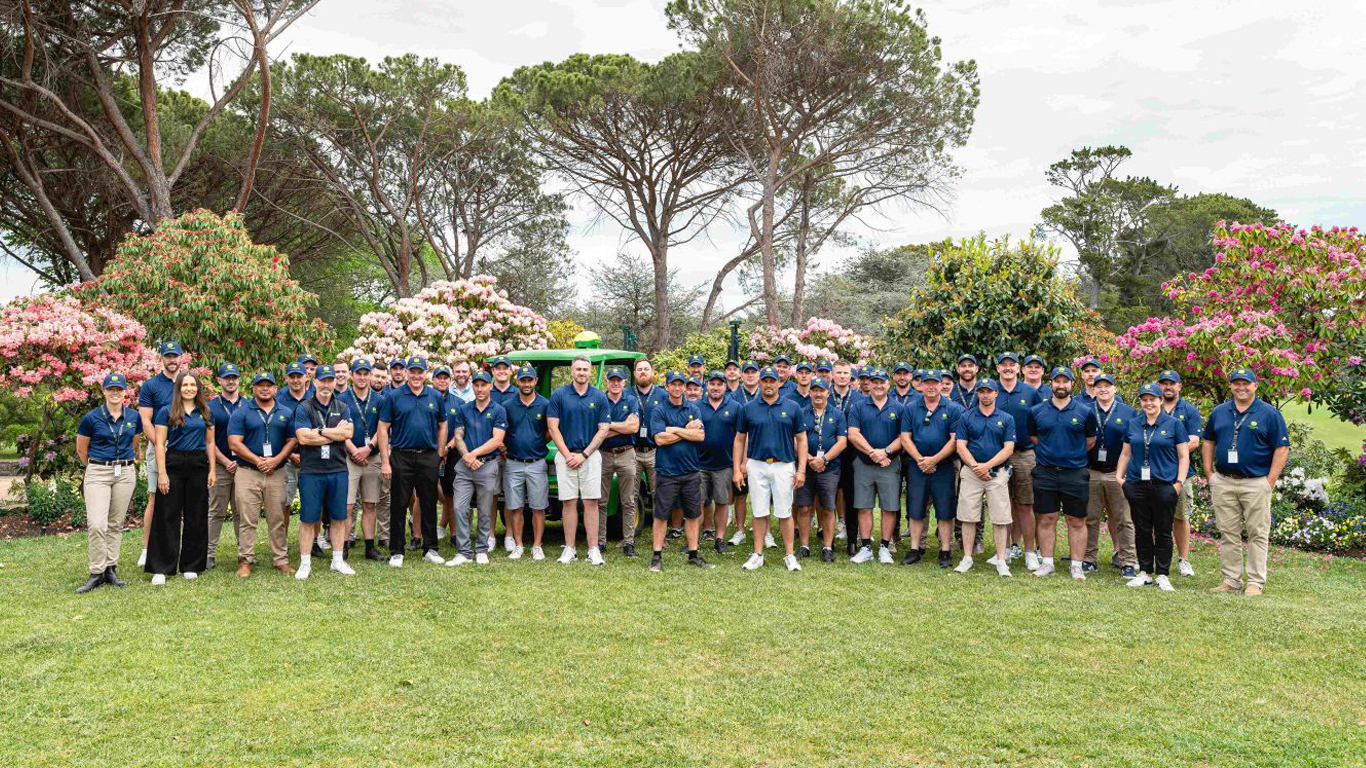 A large group of people in navy-blue polo shirts standing on a lawn with trees and shrubs behind them.