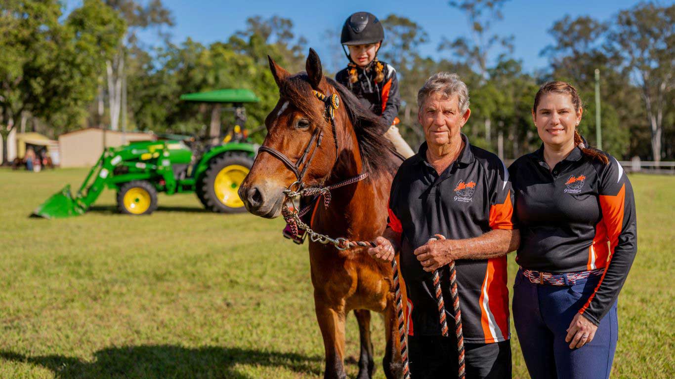 Man and woman with a bay horse and young girl in saddle, standing on grass with trees and a green tractor behind them.