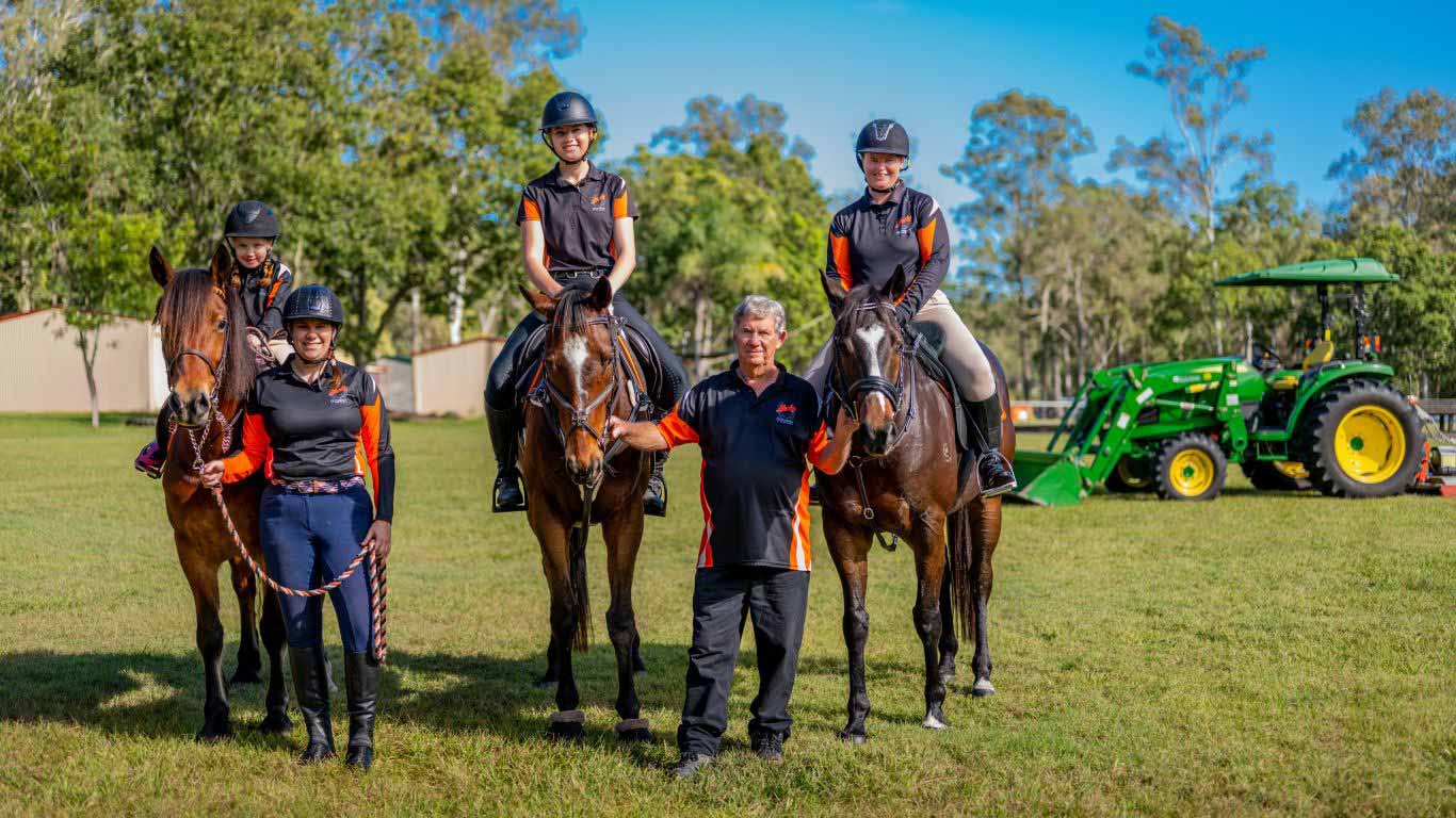 Three horses with riders are being held by a lady and a man on a green filed with sheds and trees in the background and green tractor.