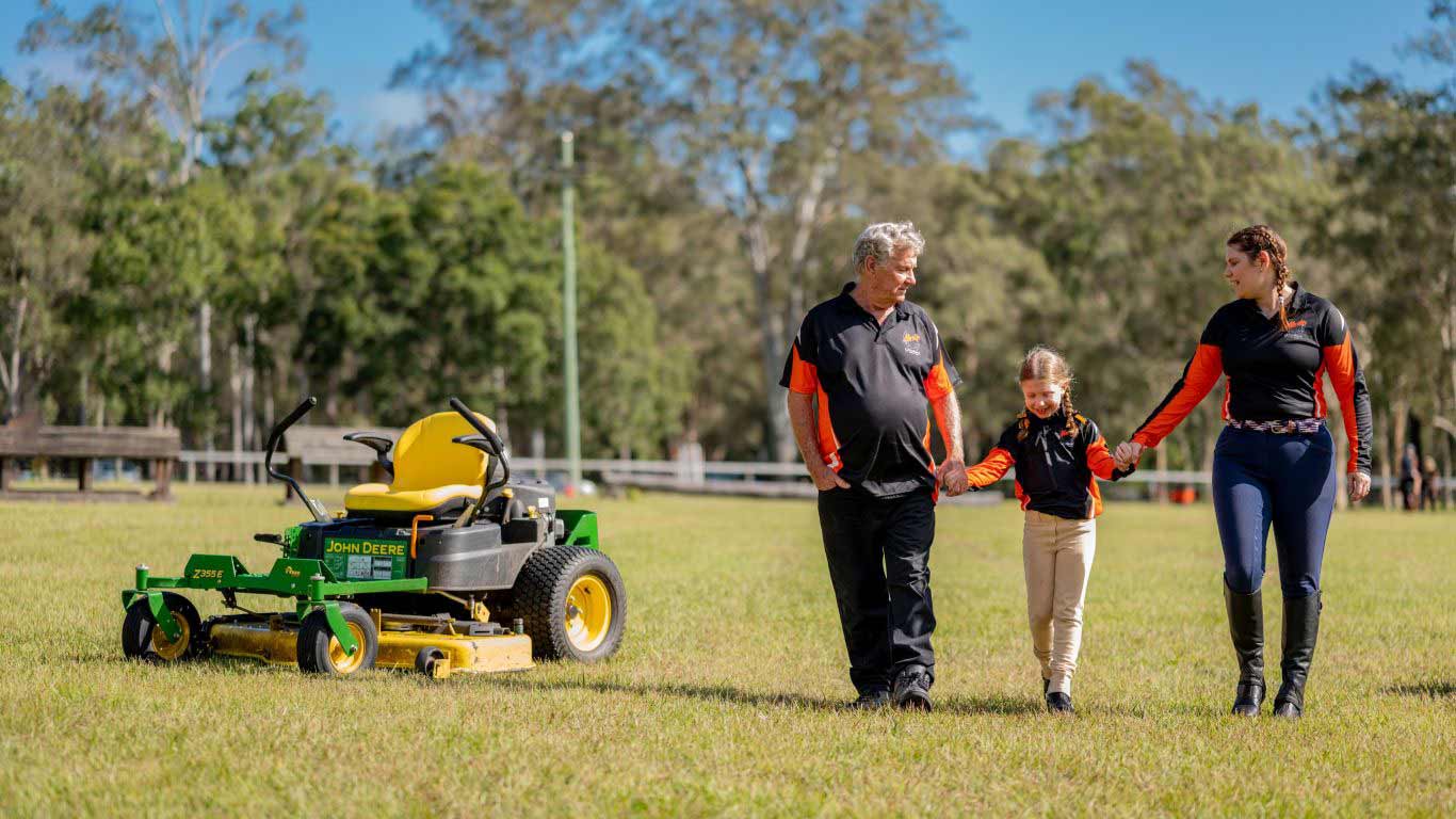Man and woman walk in a grassy field holding hands with a small girl, near a green mower with a yellow seat and trees behind.