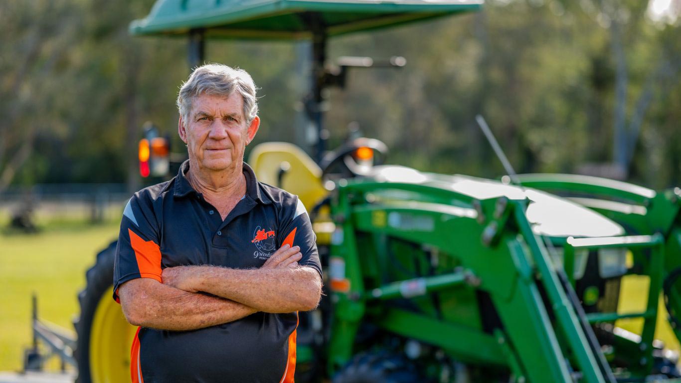 A man in a blue and orange polo shirt with his arms folded in front of a green tractor with yellow wheels.