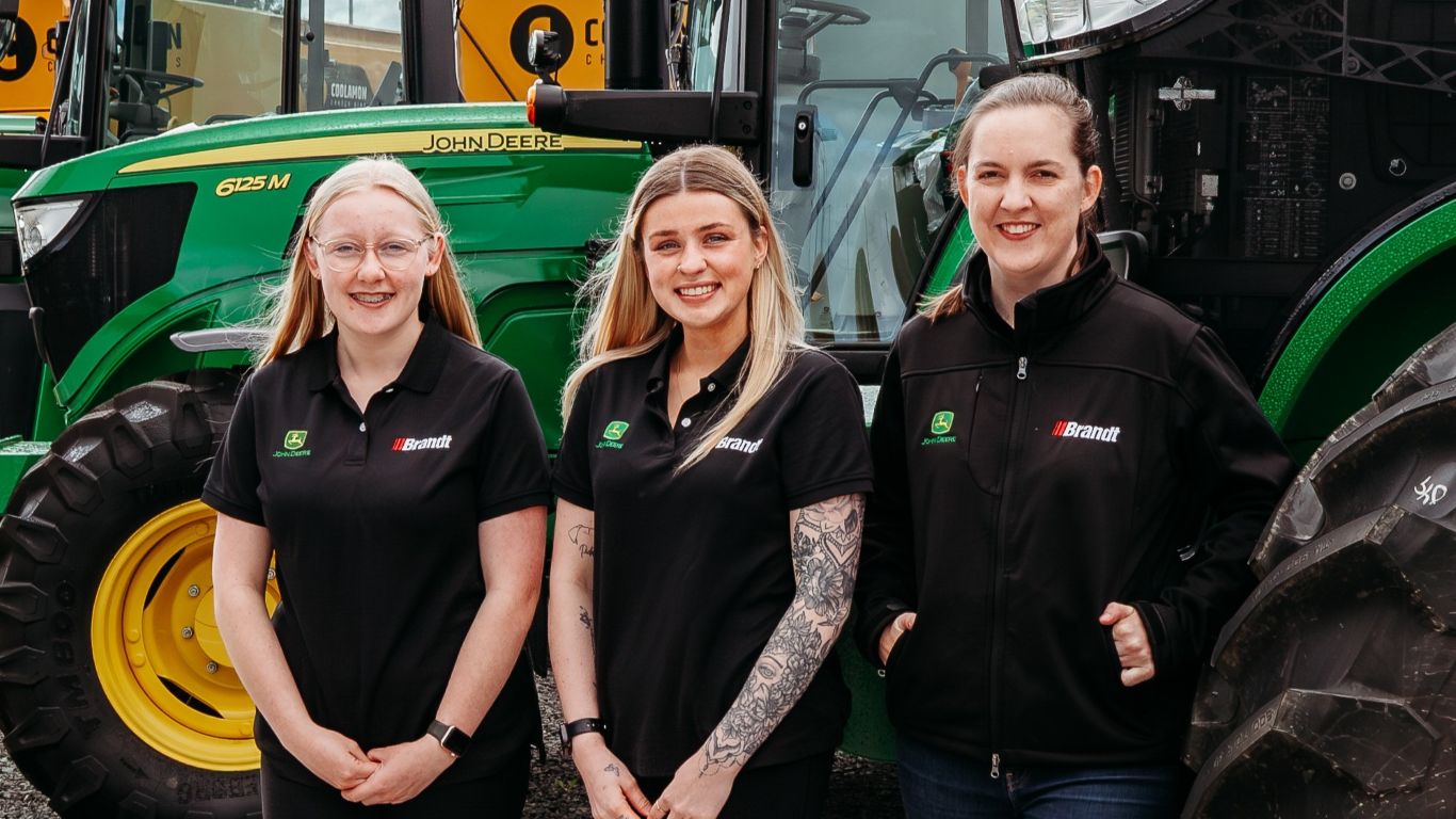 Three women in black shirts standing in line in front of green tractor outside.