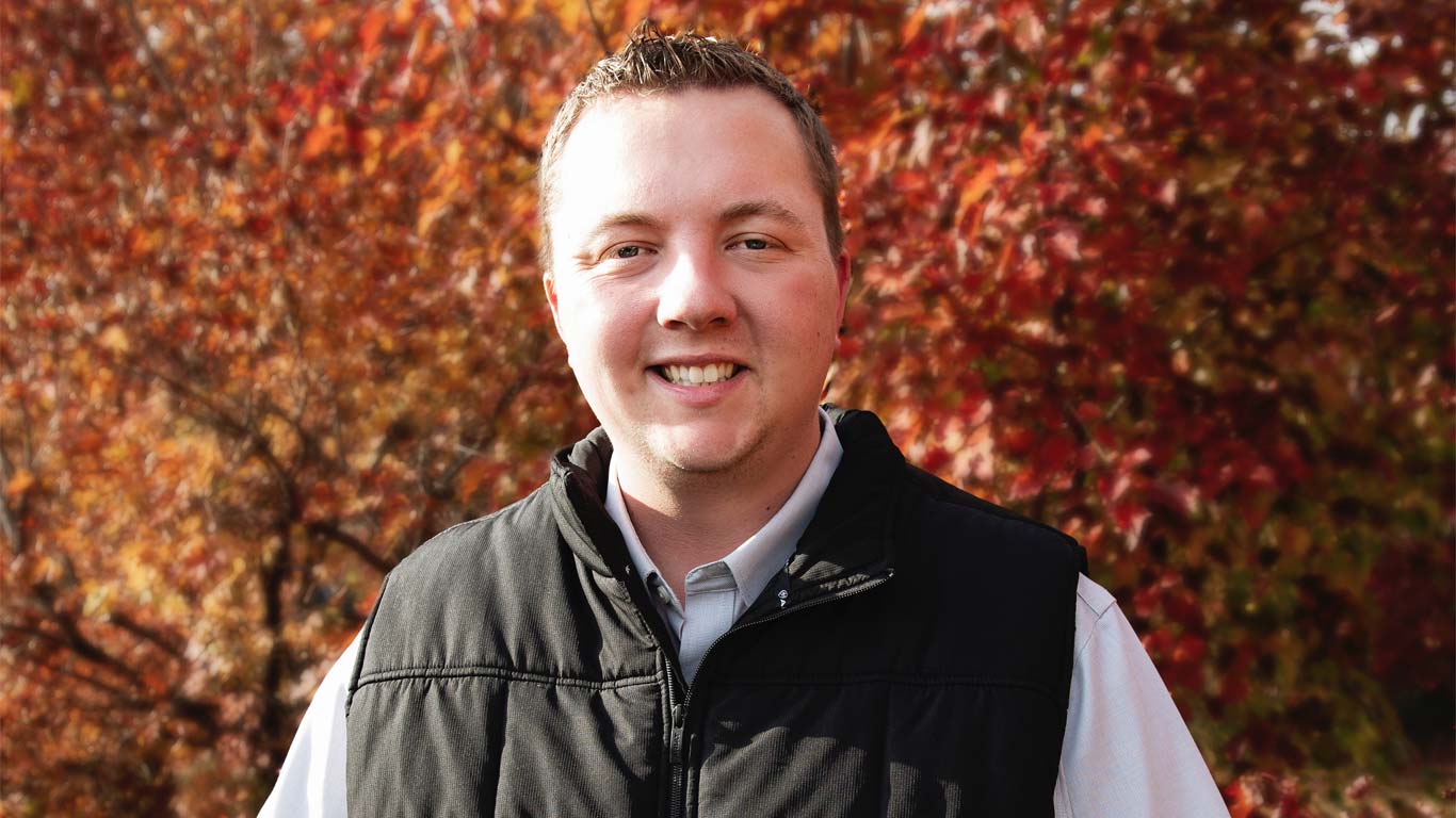 Person in a black jacket smiling outdoors with red-orange fall leaves in the background.
