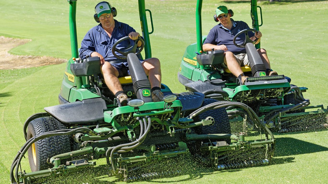 Two people in blue shirts each driving professional golf turf cutting equipment on a green lawn.