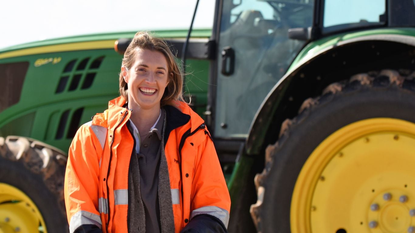 A woman in a an orange high-vis jacket standing in front of a green tractor with yellow wheels.