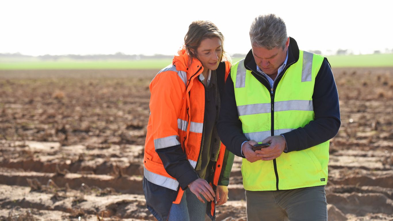 A woman in an orange high-vis jacket is looking over the shoulder of a man in a yellow high-vis vest at a phone screen.