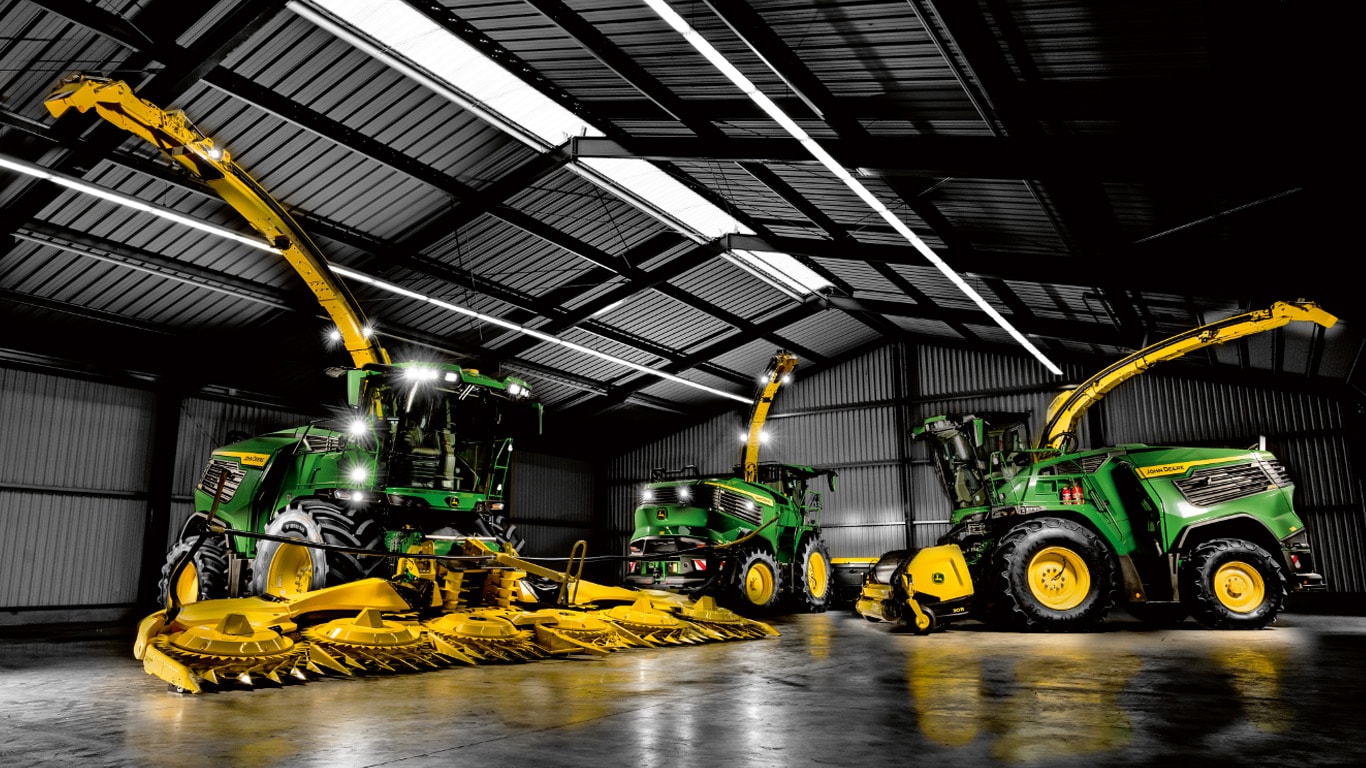 Three large forage harvesters with bright yellow headers and spouts, positioned inside a spacious, dimly lit machinery shed with a high metal roof.