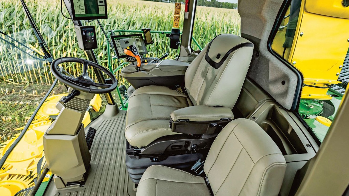 Interior view of a John Deere forage harvester cab, showing two beige leather seats, a steering wheel, multiple digital displays, and a joystick control, with a cornfield visible through the windows.